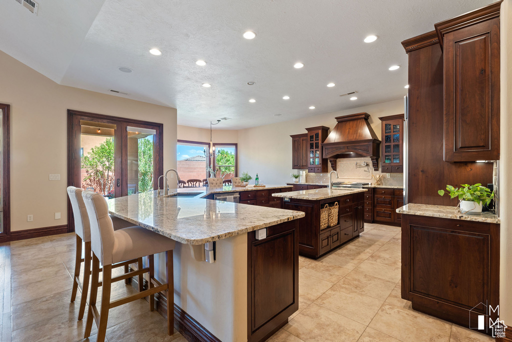 Kitchen with an island with sink, hanging light fixtures, light stone counters, a kitchen bar, and custom range hood
