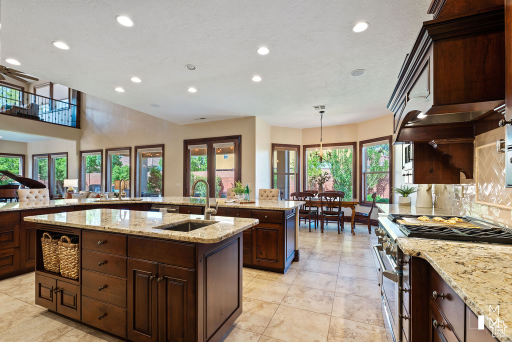 Kitchen with light stone counters, pendant lighting, light tile floors, high end stove, and a kitchen island with sink