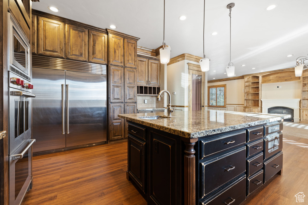 Kitchen with sink, dark hardwood / wood-style floors, light stone countertops, built in appliances, and a kitchen island with sink