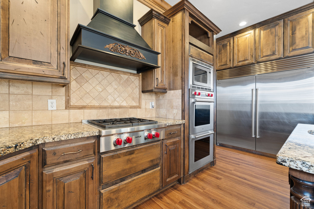 Kitchen with built in appliances, light stone counters, custom range hood, tasteful backsplash, and light hardwood / wood-style flooring