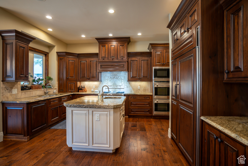 Kitchen with backsplash, built in appliances, a center island with sink, sink, and dark wood-type flooring