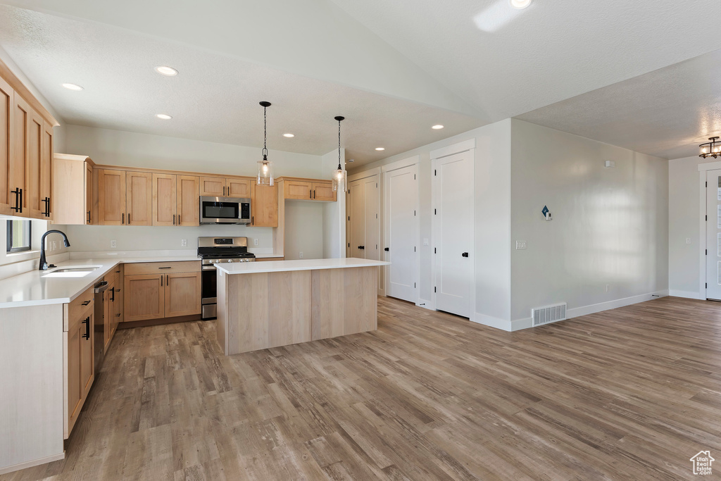 Kitchen featuring appliances with stainless steel finishes, light wood-type flooring, a center island, decorative light fixtures, and light brown cabinetry