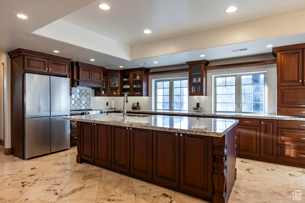 Kitchen featuring backsplash, a kitchen island with sink, light stone counters, sink, and stainless steel fridge