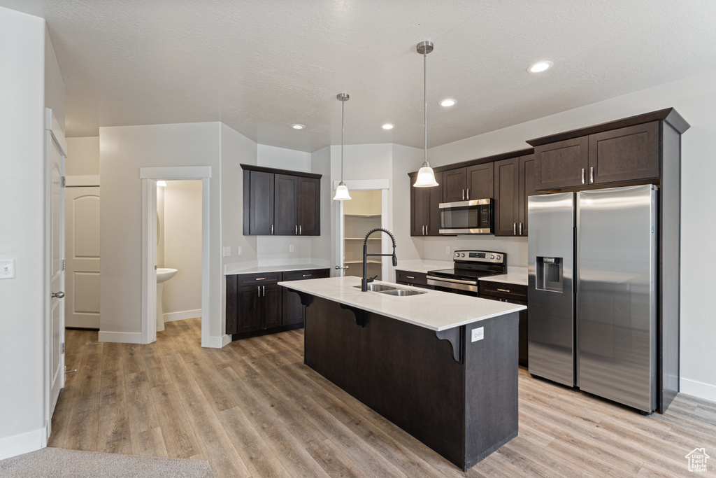 Kitchen with appliances with stainless steel finishes, hanging light fixtures, light hardwood / wood-style flooring, sink, and a center island with sink