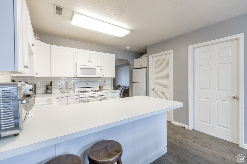 Kitchen featuring white cabinets, a breakfast bar area, white appliances, tasteful backsplash, and dark wood-type flooring