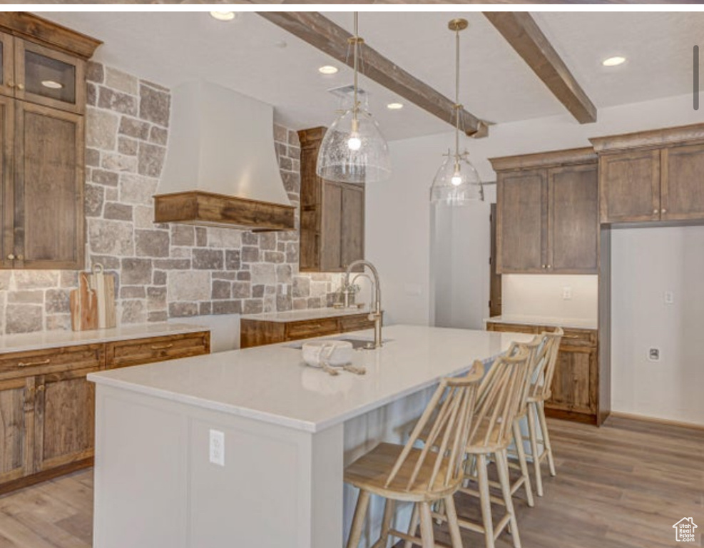 Kitchen featuring pendant lighting, light hardwood / wood-style flooring, custom range hood, beamed ceiling, and a kitchen island with sink