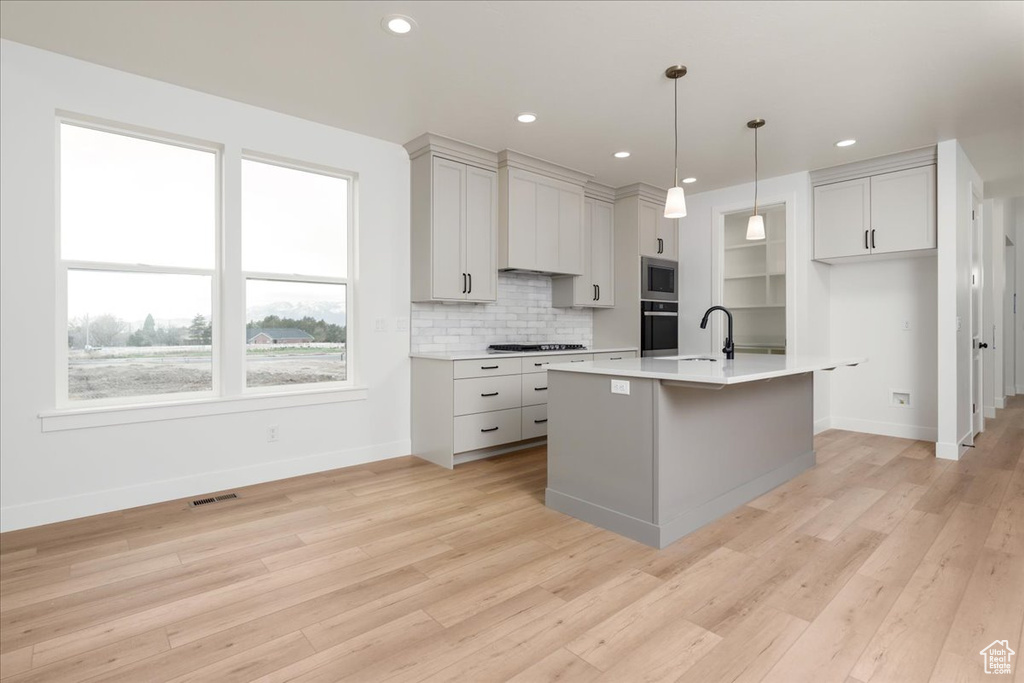 Kitchen featuring appliances with stainless steel finishes, an island with sink, light wood-type flooring, tasteful backsplash, and decorative light fixtures