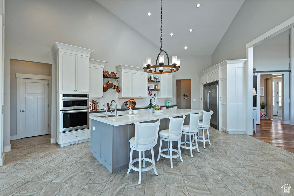 Kitchen with light wood-type flooring, a center island with sink, stainless steel appliances, white cabinetry, and high vaulted ceiling