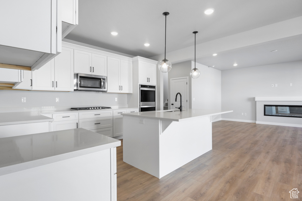 Kitchen featuring appliances with stainless steel finishes, white cabinets, an island with sink, and decorative light fixtures