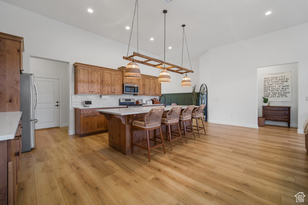 Kitchen with light hardwood / wood-style flooring, a center island with sink, hanging light fixtures, appliances with stainless steel finishes, and tasteful backsplash