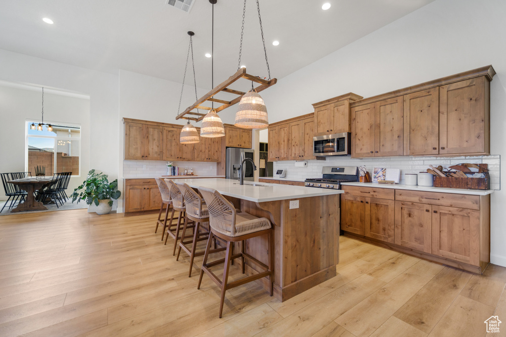 Kitchen featuring a kitchen island with sink, light wood-type flooring, hanging light fixtures, appliances with stainless steel finishes, and tasteful backsplash