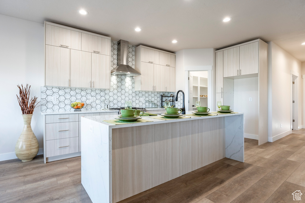 Kitchen with light hardwood / wood-style flooring, wall chimney range hood, backsplash, and a kitchen island with sink