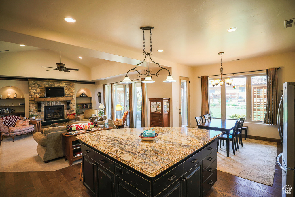 Kitchen with light stone counters, a stone fireplace, hanging light fixtures, a center island, and ceiling fan with notable chandelier