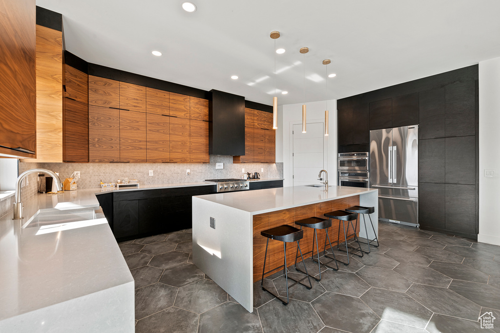 Kitchen featuring backsplash, appliances with stainless steel finishes, a kitchen bar, and a center island with sink
