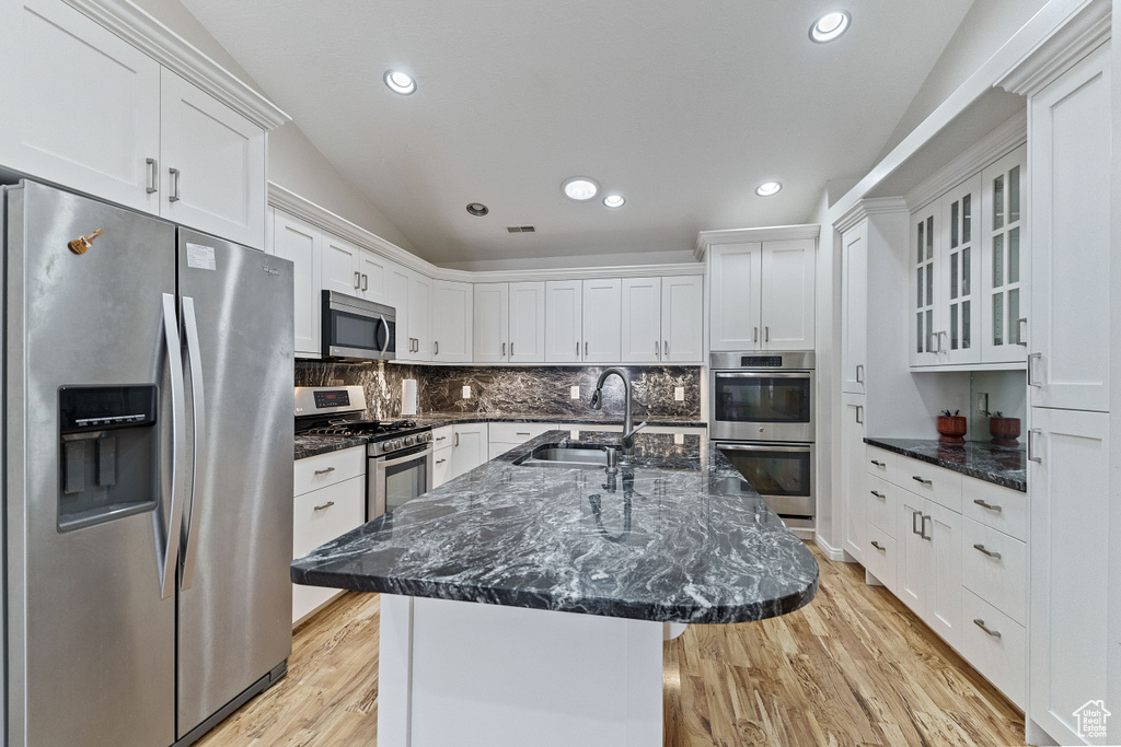 Kitchen with backsplash, a kitchen island with sink, stainless steel appliances, white cabinetry, and light hardwood / wood-style flooring