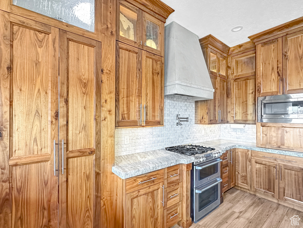 Kitchen featuring light stone countertops, light hardwood / wood-style floors, stainless steel appliances, premium range hood, and backsplash