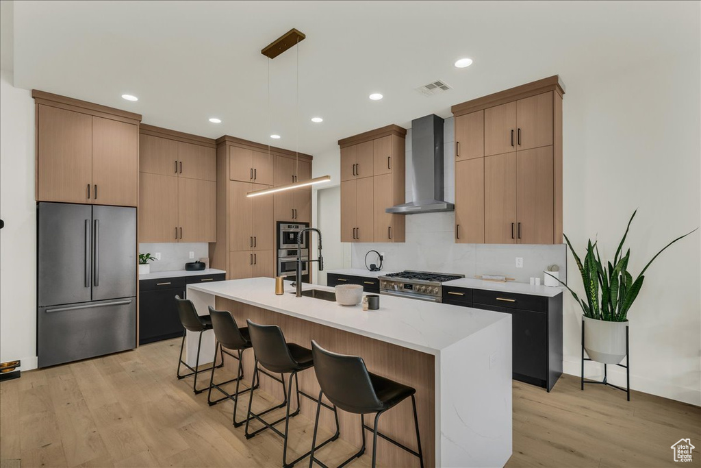 Kitchen with a kitchen island with sink, hanging light fixtures, wall chimney exhaust hood, high quality appliances, and light wood-type flooring