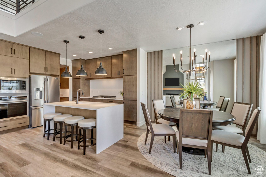 Kitchen with hanging light fixtures, an inviting chandelier, appliances with stainless steel finishes, an island with sink, and light wood-type flooring