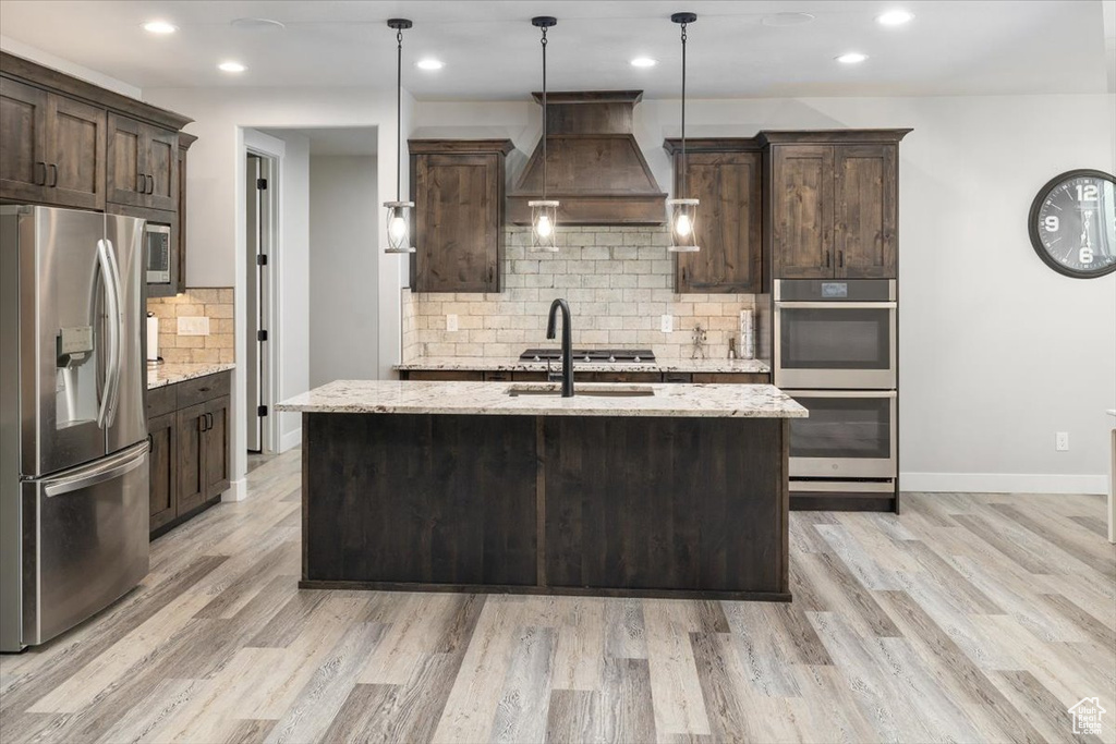 Kitchen featuring premium range hood, light hardwood / wood-style floors, light stone countertops, tasteful backsplash, and appliances with stainless steel finishes