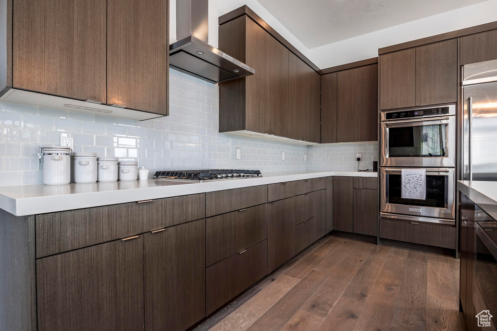 Kitchen featuring dark hardwood / wood-style floors, appliances with stainless steel finishes, wall chimney exhaust hood, tasteful backsplash, and dark brown cabinetry