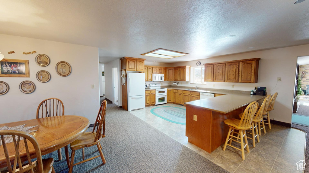 Kitchen with kitchen peninsula, white appliances, light tile floors, a kitchen bar, and a textured ceiling