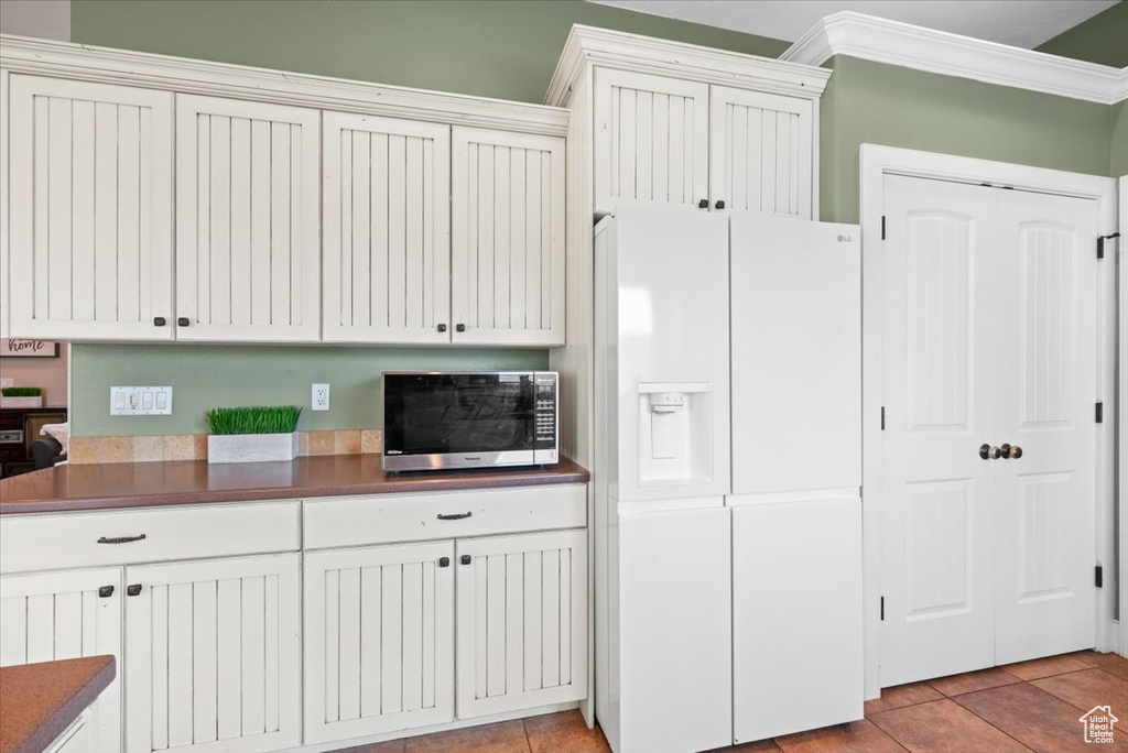 Kitchen featuring white fridge with ice dispenser, ornamental molding, light tile flooring, and white cabinetry