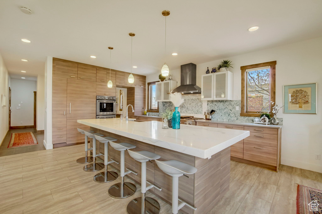 Kitchen featuring a kitchen breakfast bar, hanging light fixtures, a center island with sink, light stone countertops, and wall chimney range hood