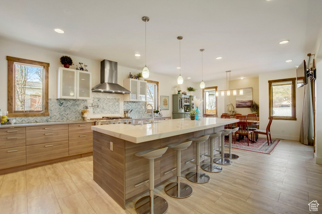 Kitchen featuring decorative light fixtures, backsplash, a kitchen island with sink, a kitchen breakfast bar, and wall chimney range hood