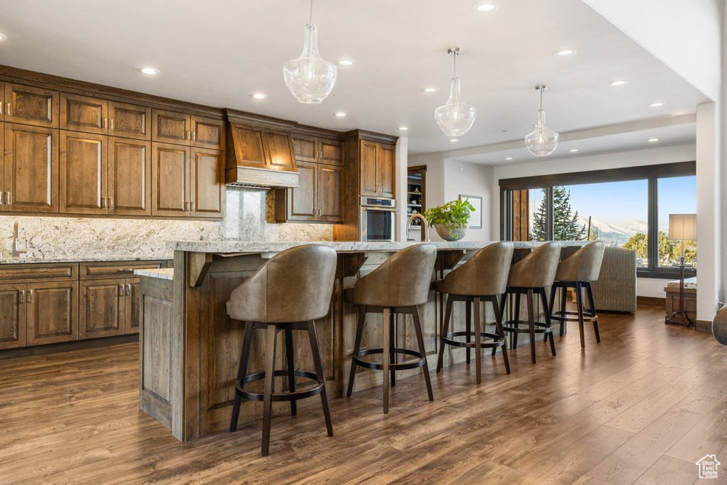 Kitchen featuring an island with sink, stainless steel oven, hanging light fixtures, custom range hood, and dark hardwood / wood-style floors