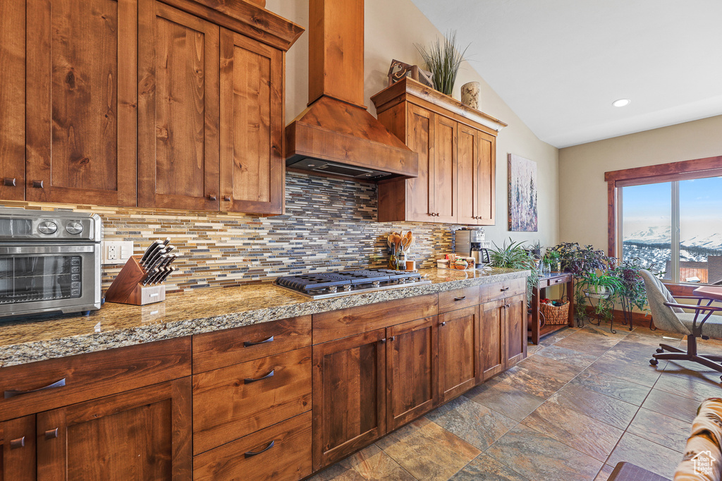 Kitchen featuring light stone counters, vaulted ceiling, dark tile floors, tasteful backsplash, and custom range hood