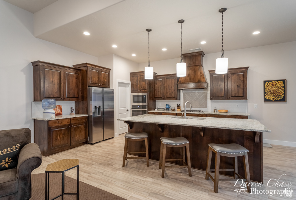 Kitchen featuring appliances with stainless steel finishes, light hardwood / wood-style flooring, custom range hood, and a center island with sink