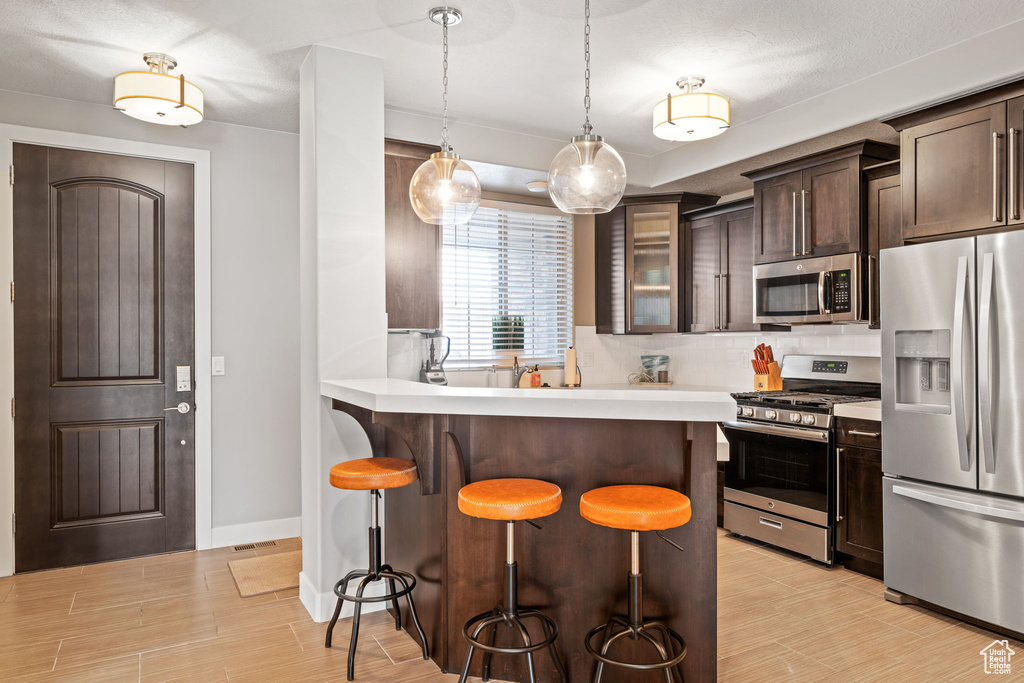 Kitchen featuring hanging light fixtures, a kitchen bar, stainless steel appliances, dark brown cabinets, and tasteful backsplash