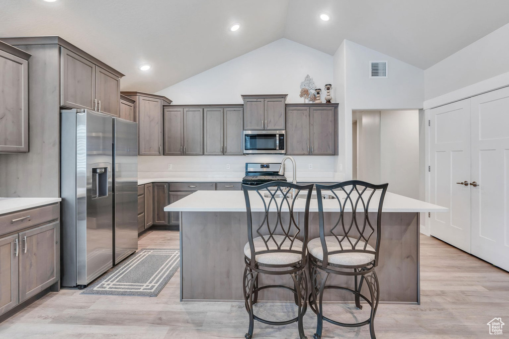 Kitchen featuring a breakfast bar, high vaulted ceiling, light hardwood / wood-style flooring, stainless steel appliances, and a center island with sink