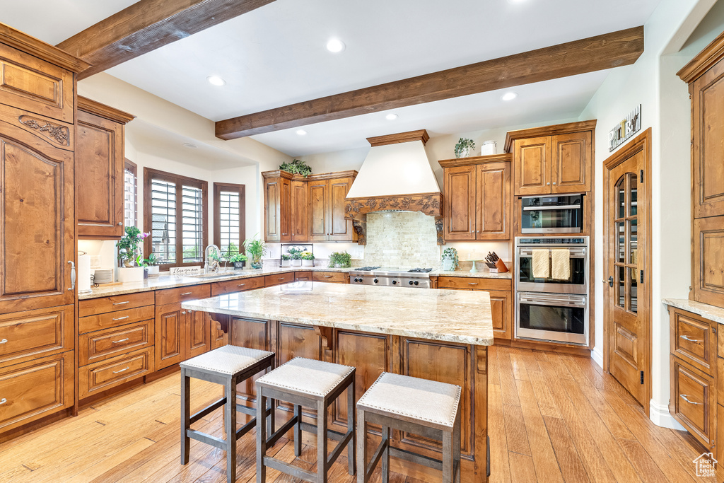 Kitchen featuring premium range hood, light hardwood / wood-style floors, a center island, beam ceiling, and light stone counters