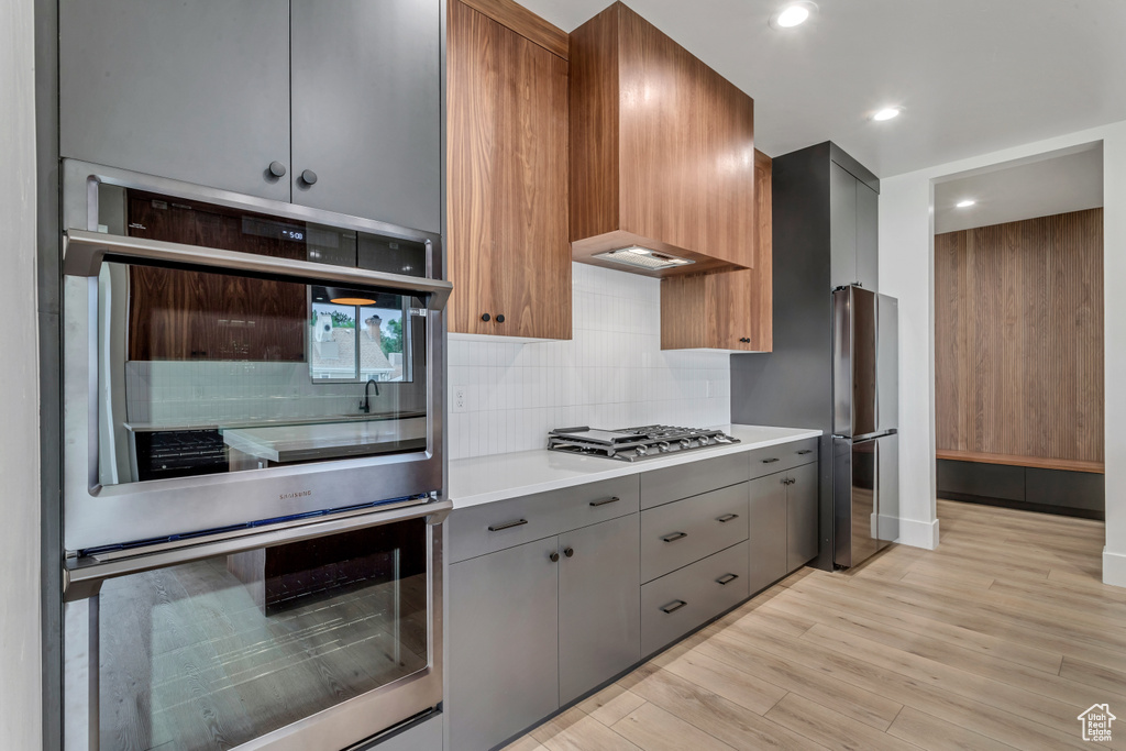 Kitchen with gray cabinets, appliances with stainless steel finishes, backsplash, light hardwood / wood-style floors, and premium range hood