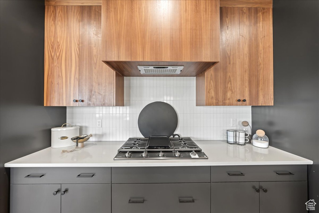 Kitchen with exhaust hood, premium range hood, stainless steel gas stovetop, gray cabinets, and backsplash