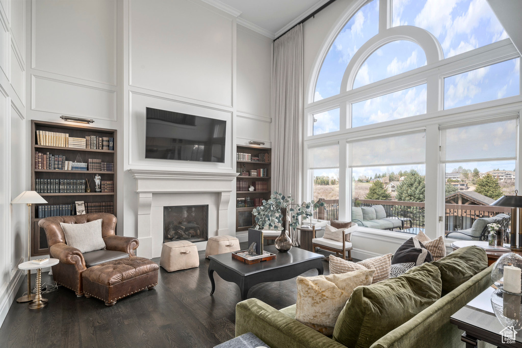 Living room featuring built in shelves, dark wood-type flooring, a towering ceiling, and crown molding