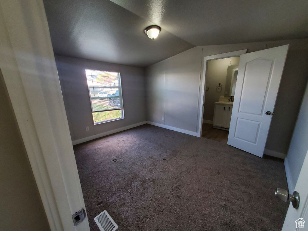 Unfurnished bedroom with ensuite bath, vaulted ceiling, and dark carpet