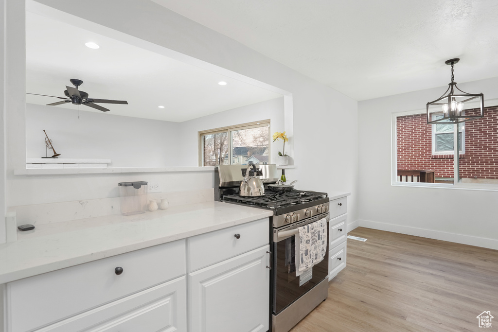 Kitchen featuring stainless steel range with gas cooktop, ceiling fan with notable chandelier, light hardwood / wood-style flooring, white cabinets, and pendant lighting