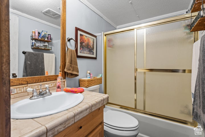 Full bathroom with toilet, a textured ceiling, vanity, ornamental molding, and combined bath / shower with glass door