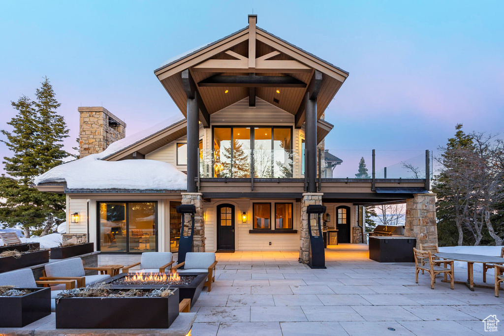 Back house at dusk featuring a balcony, an outdoor fire pit, exterior kitchen, and a patio