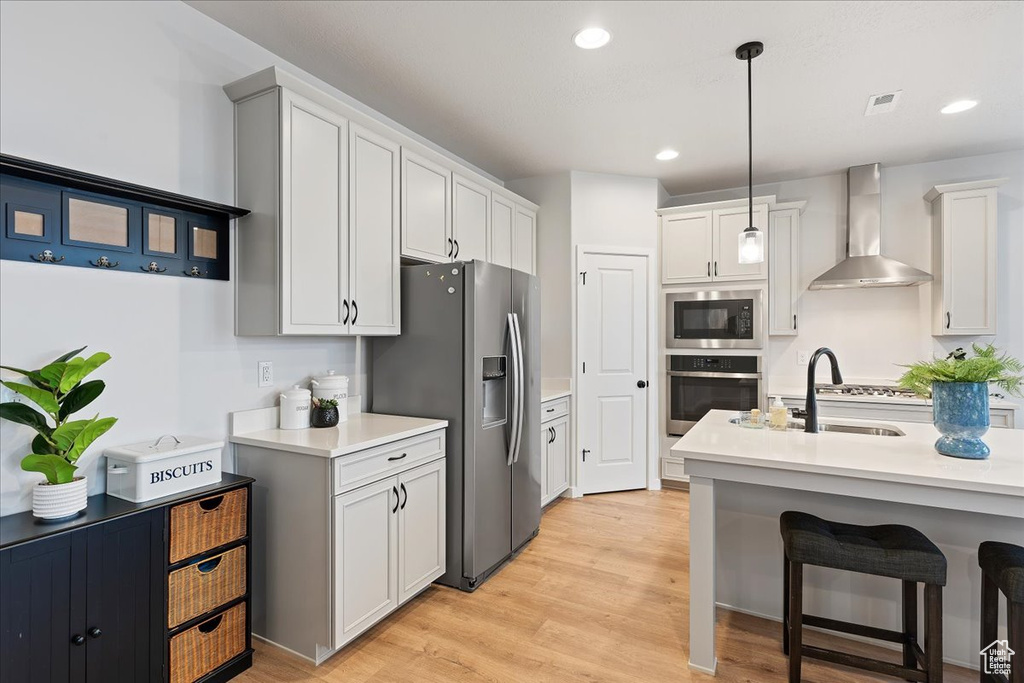 Kitchen with decorative light fixtures, appliances with stainless steel finishes, light hardwood / wood-style floors, white cabinets, and wall chimney range hood