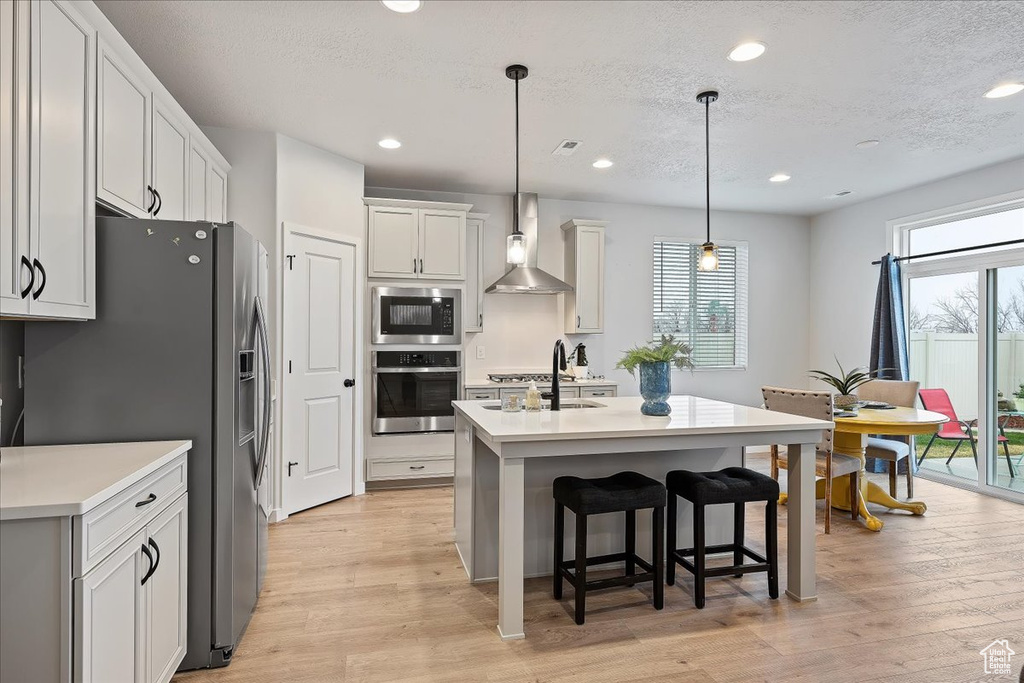 Kitchen featuring appliances with stainless steel finishes, light wood-type flooring, a center island with sink, wall chimney range hood, and pendant lighting
