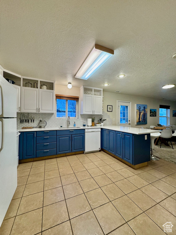 Kitchen with kitchen peninsula, white appliances, blue cabinets, light tile floors, and white cabinets