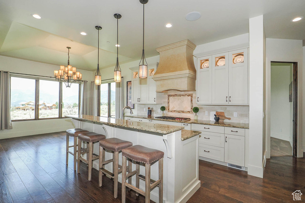 Kitchen featuring a chandelier, tasteful backsplash, custom range hood, a kitchen island with sink, and light stone counters