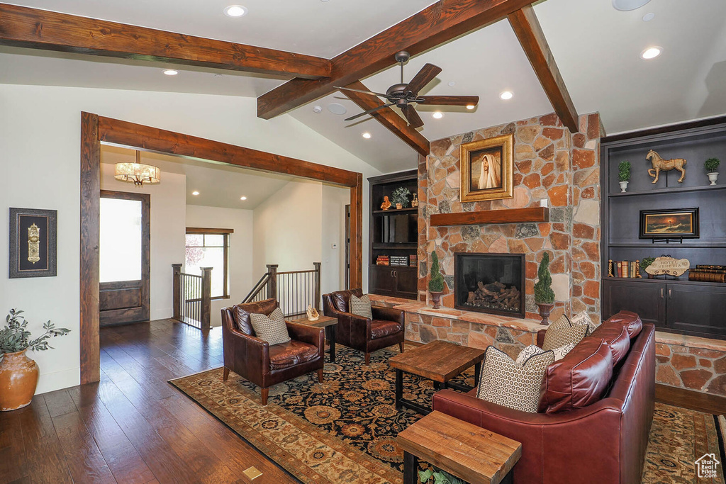 Living room featuring vaulted ceiling with beams, ceiling fan with notable chandelier, a stone fireplace, dark wood-type flooring, and built in shelves