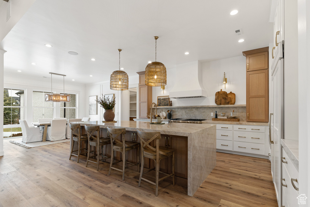Kitchen featuring custom range hood, a breakfast bar area, a center island with sink, tasteful backsplash, and light wood-type flooring