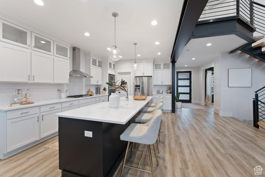 Kitchen featuring appliances with stainless steel finishes, a center island with sink, wall chimney exhaust hood, decorative light fixtures, and light wood-type flooring