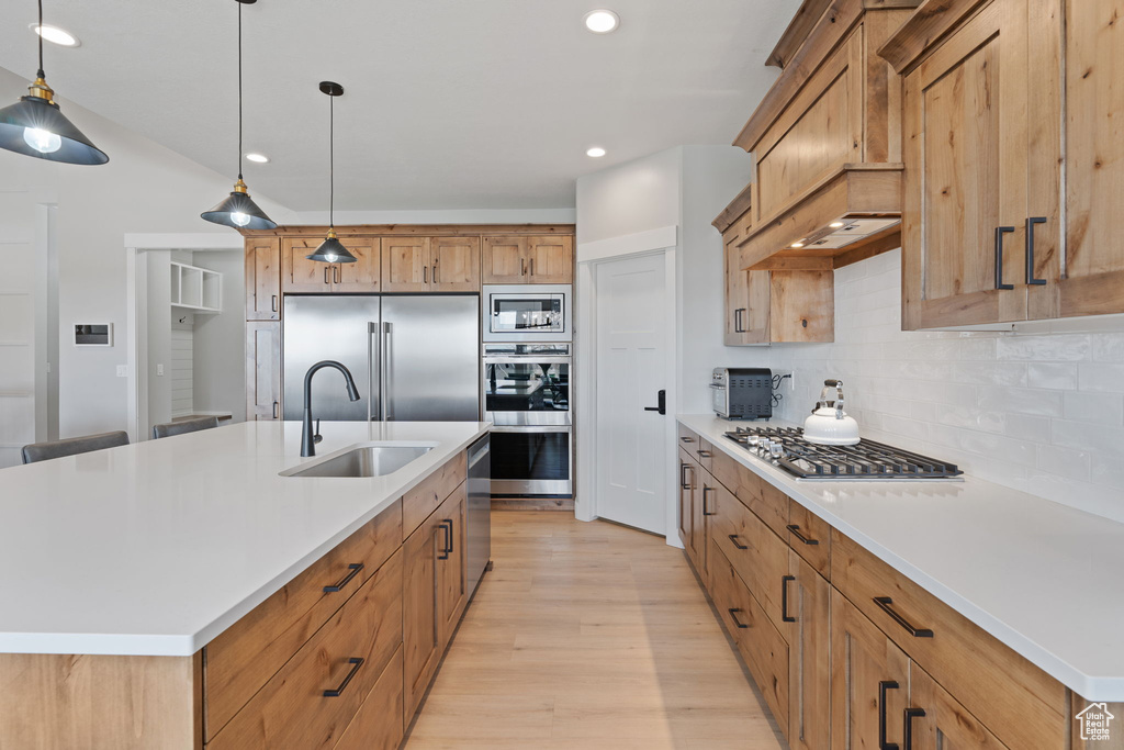 Kitchen featuring decorative light fixtures, light hardwood / wood-style flooring, an island with sink, tasteful backsplash, and built in appliances