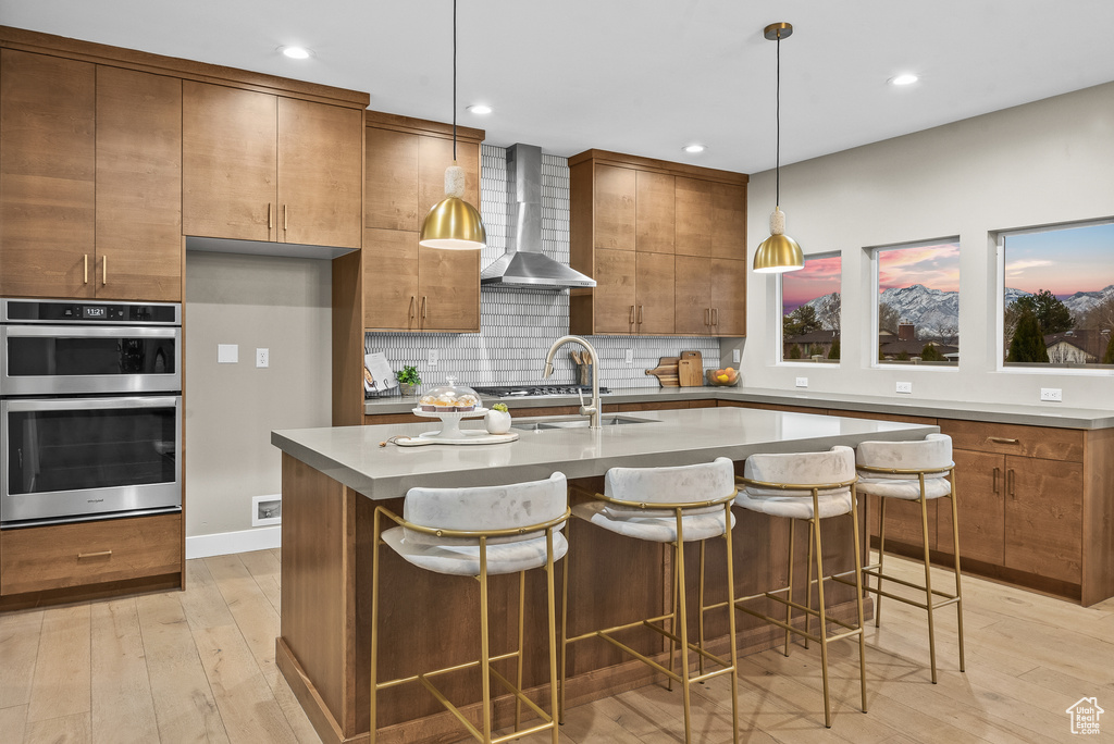 Kitchen featuring a kitchen island with sink, stainless steel double oven, wall chimney exhaust hood, decorative light fixtures, and light wood-type flooring